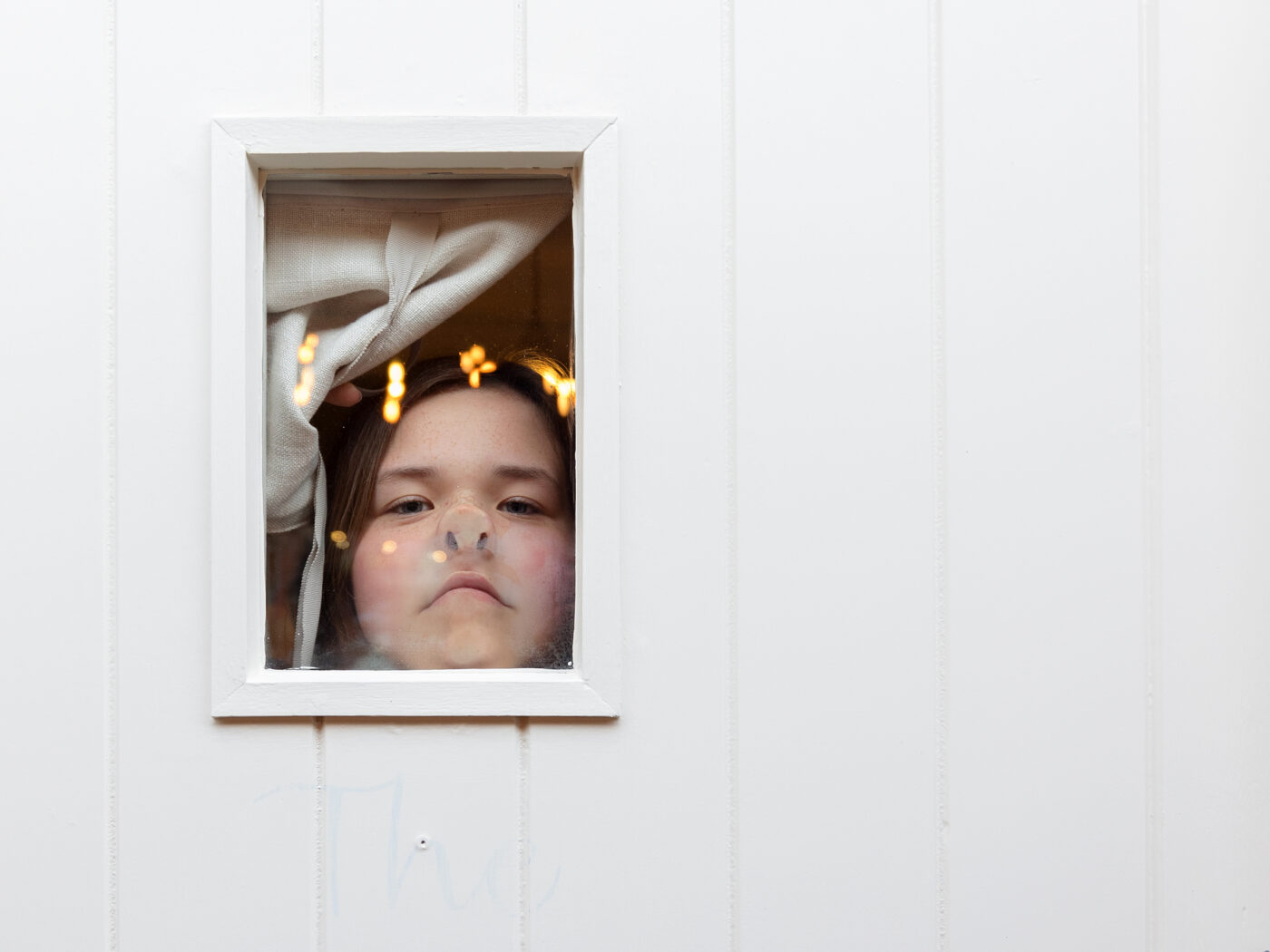 A girl is looking through a window in a door. Her face is pressed up against the glass pushing her nose up. She is looking straight at the camera. The photo was taken at The Old Cow shed during a wedding