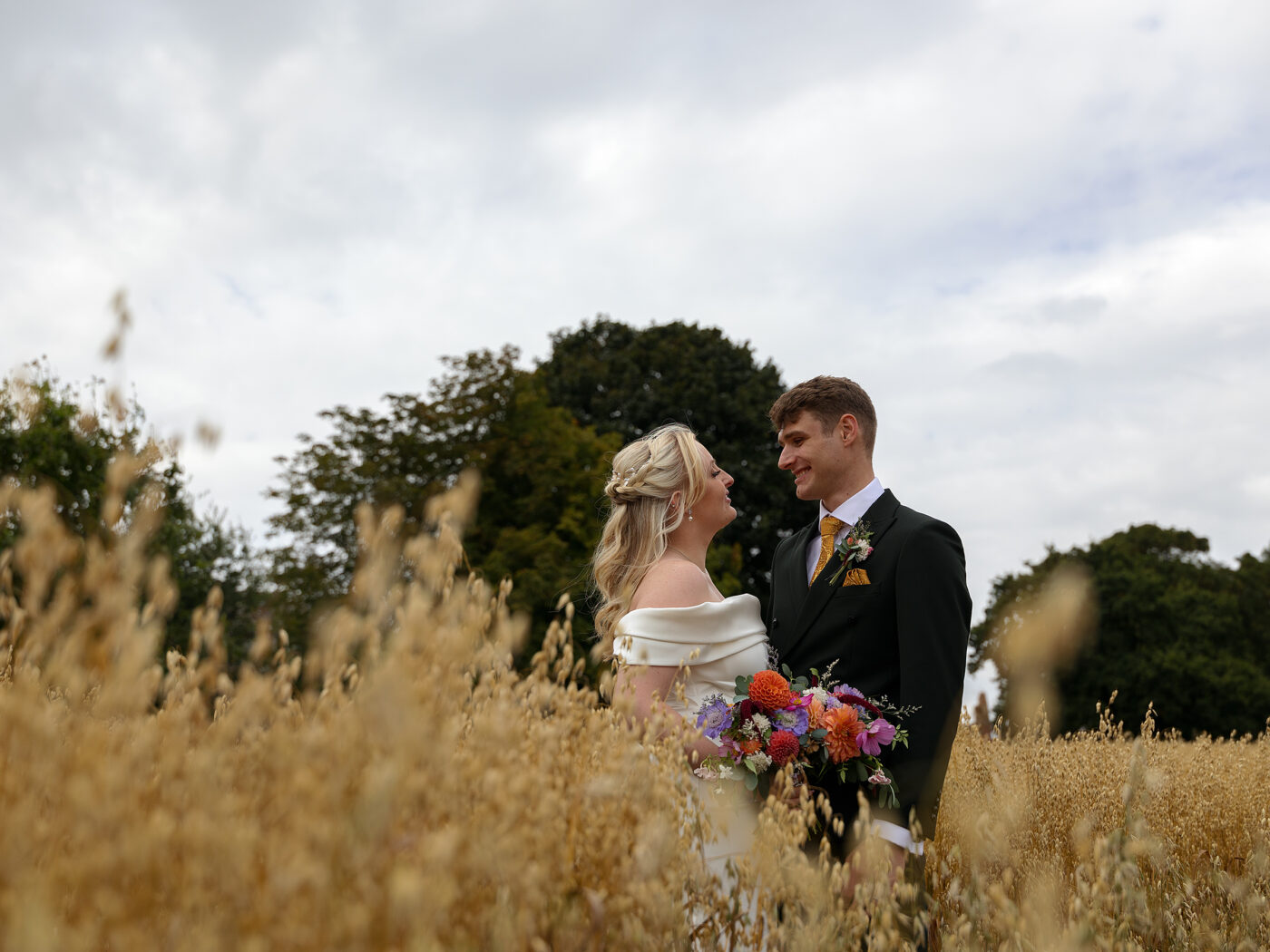 bride and groom stood in a wheat field, side by side looking into each others eyes.