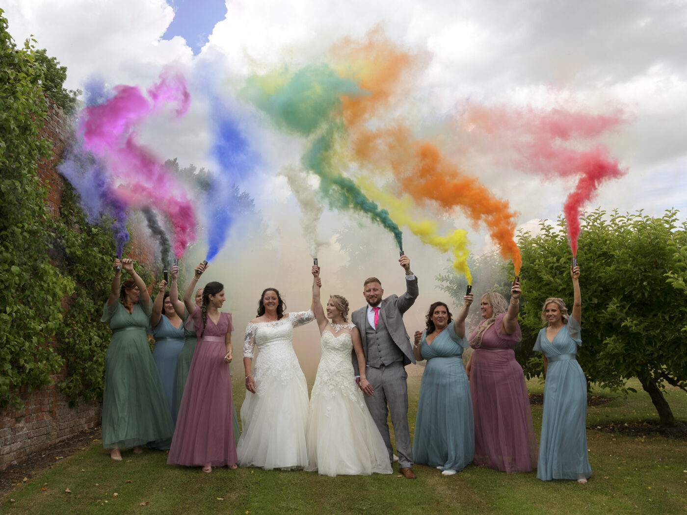 2 brides and their wedding party are walking in a row, holding different coloured smoke bombs in the air