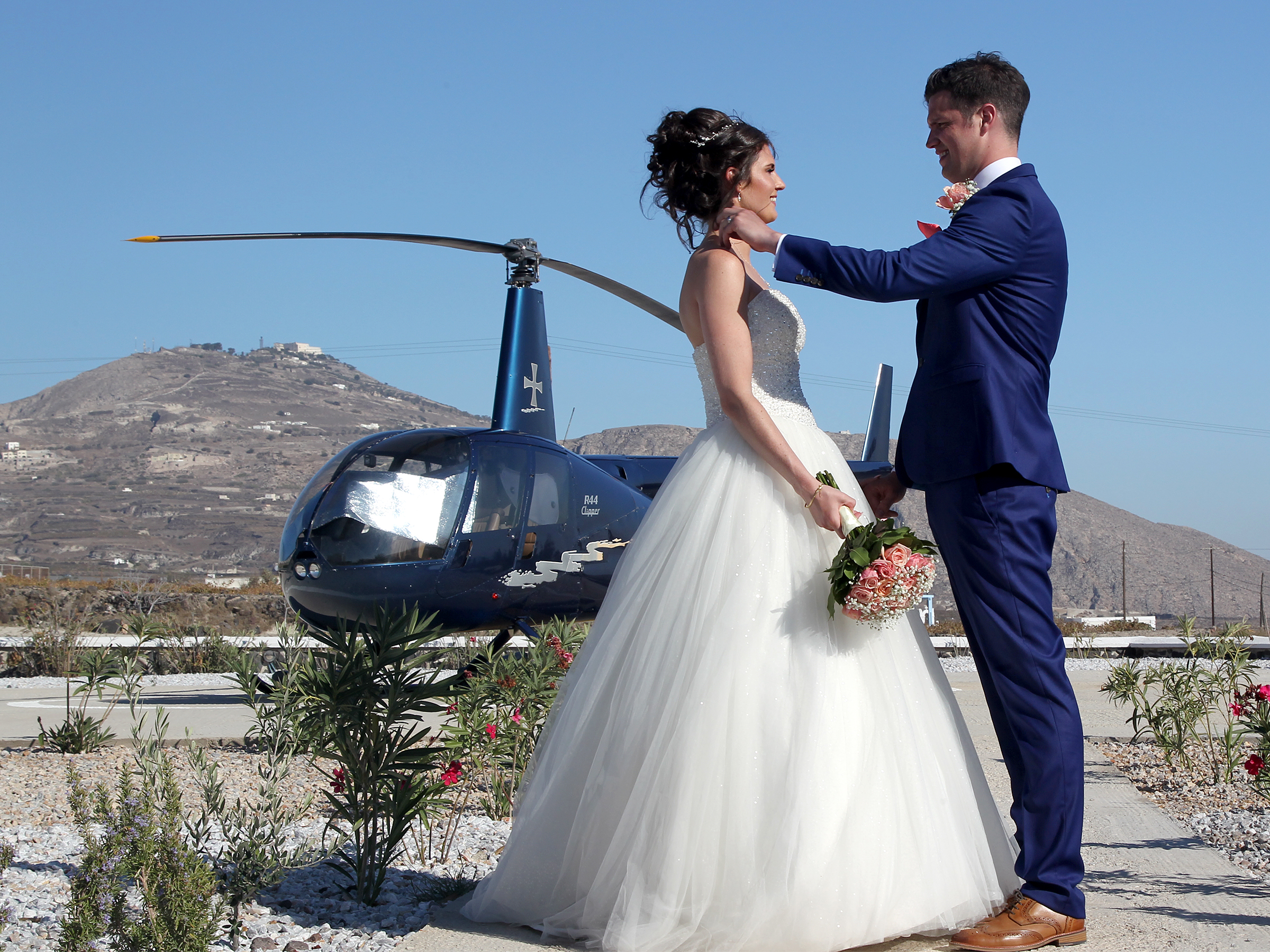 Bride and groom stood facing each other, the groom is tucking the brides hair behind her ear. There is a helicopter behind them.