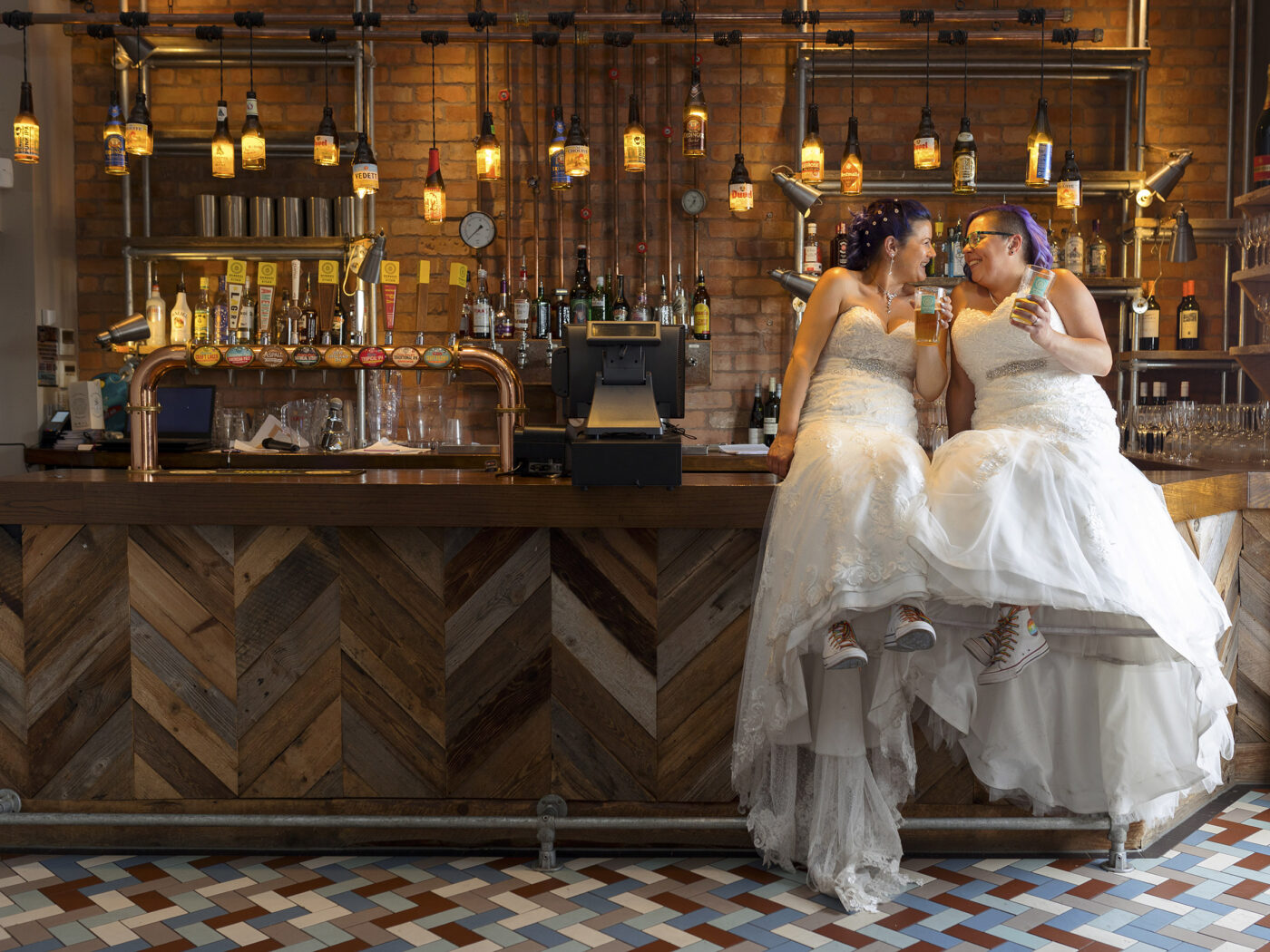 Nottingham wedding blog showing 2 brides are sitting on the end of a pub bar. There are lots of lights hanging behind them. They are both wearing wedding dresses and rainbow soled white converse. They both have a pint in their hands.