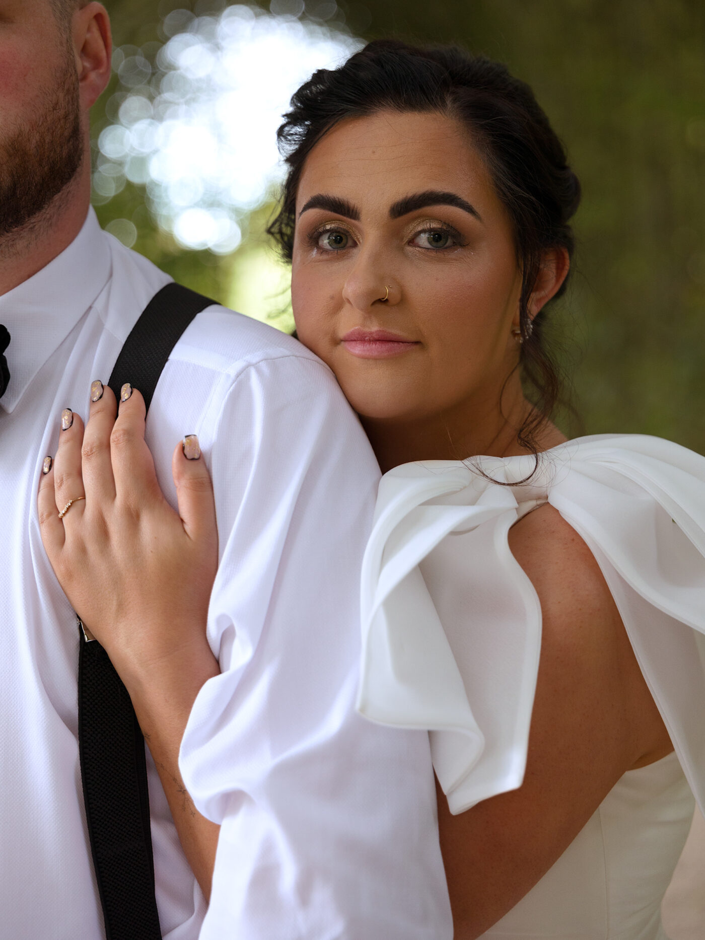 Dark haired Bride wearing a Vera wang wedding dress with oversized bow on her shoulder hugs her new husband from behind and looks directly into the camera. The wedding took place at Alnwick Tree House and the photographs were taken in Alnwick gardens