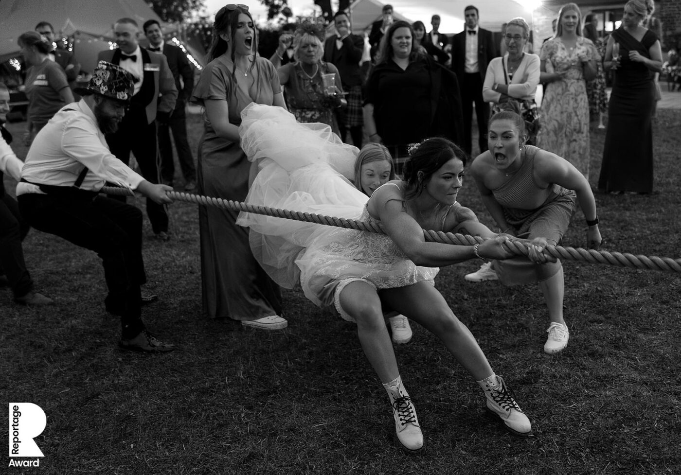 Bride and groom playfully compete in a tug-of-war game with guests cheering behind them during a lively outdoor wedding reception. This is reportage image winner