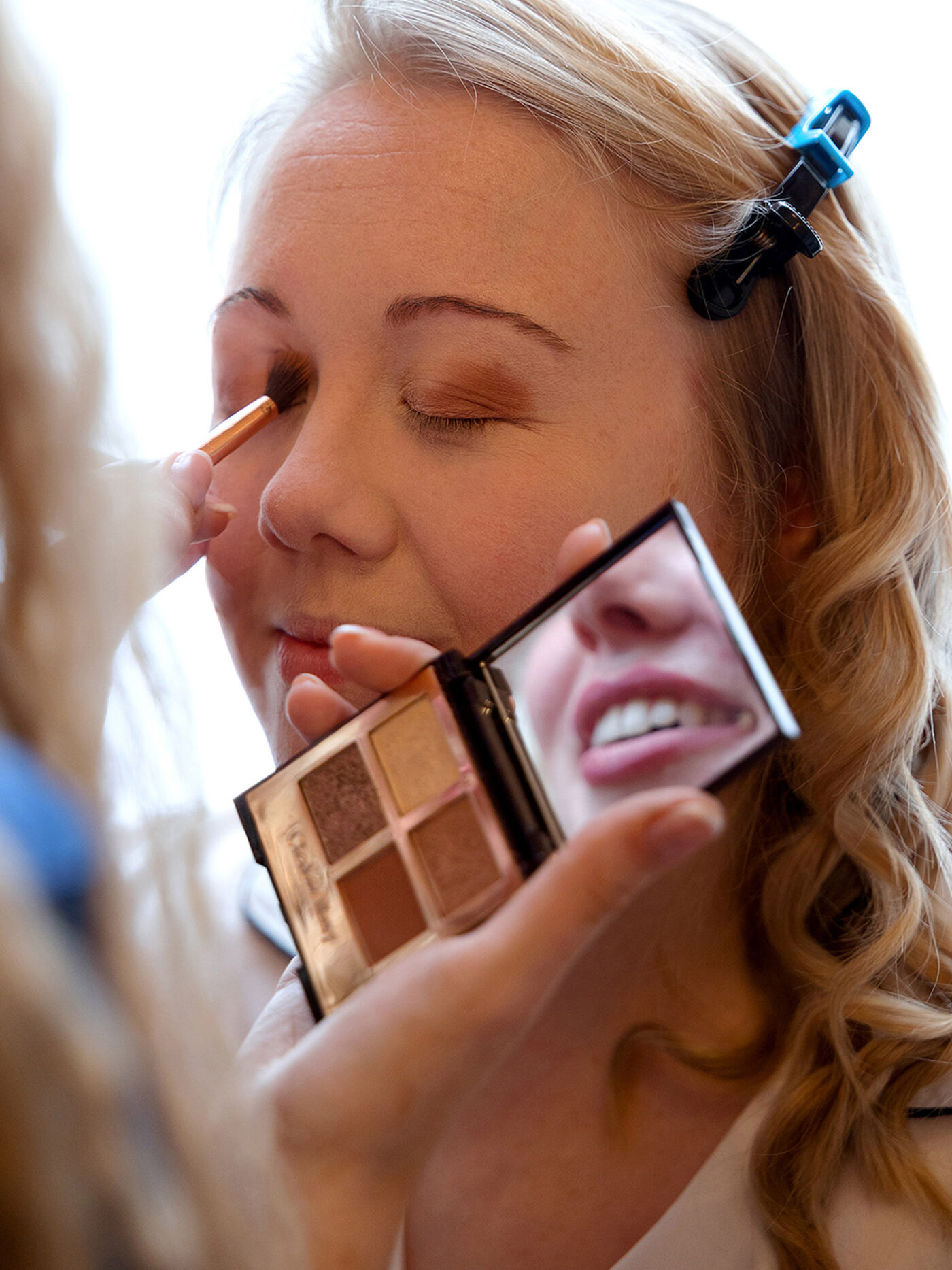 A Bride is having her make up done. The MUAs smile is reflected in a hand held mirror