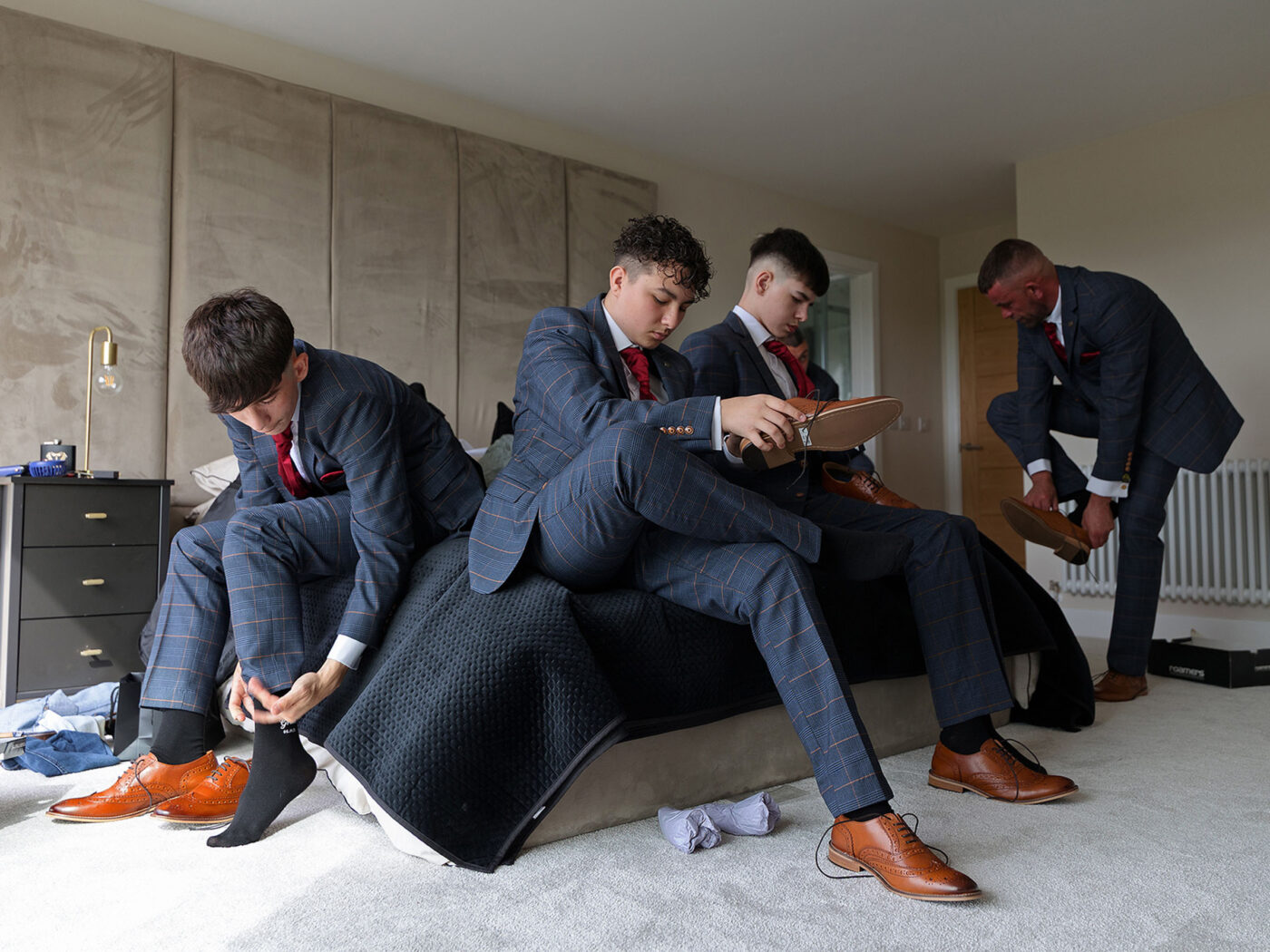 3 suited young men are sitting on the edge of a bed putting their shoes on.