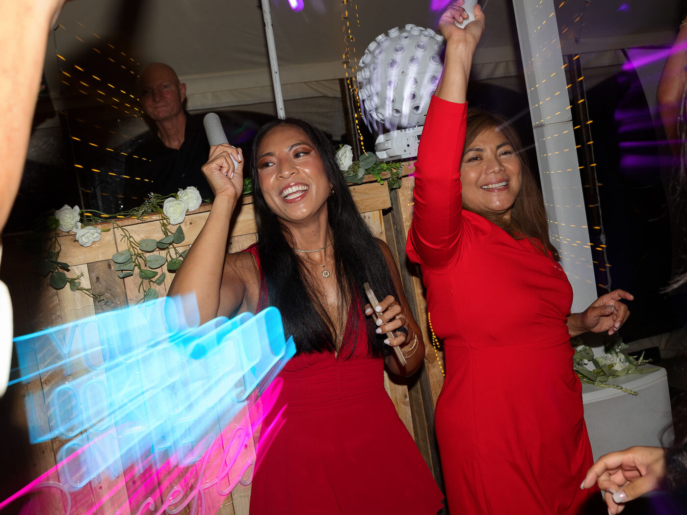 2 women are dancing at a wedding, with their arms in the air. Both are wearing red dresses on. The colour red at a wedding is culturally traditional