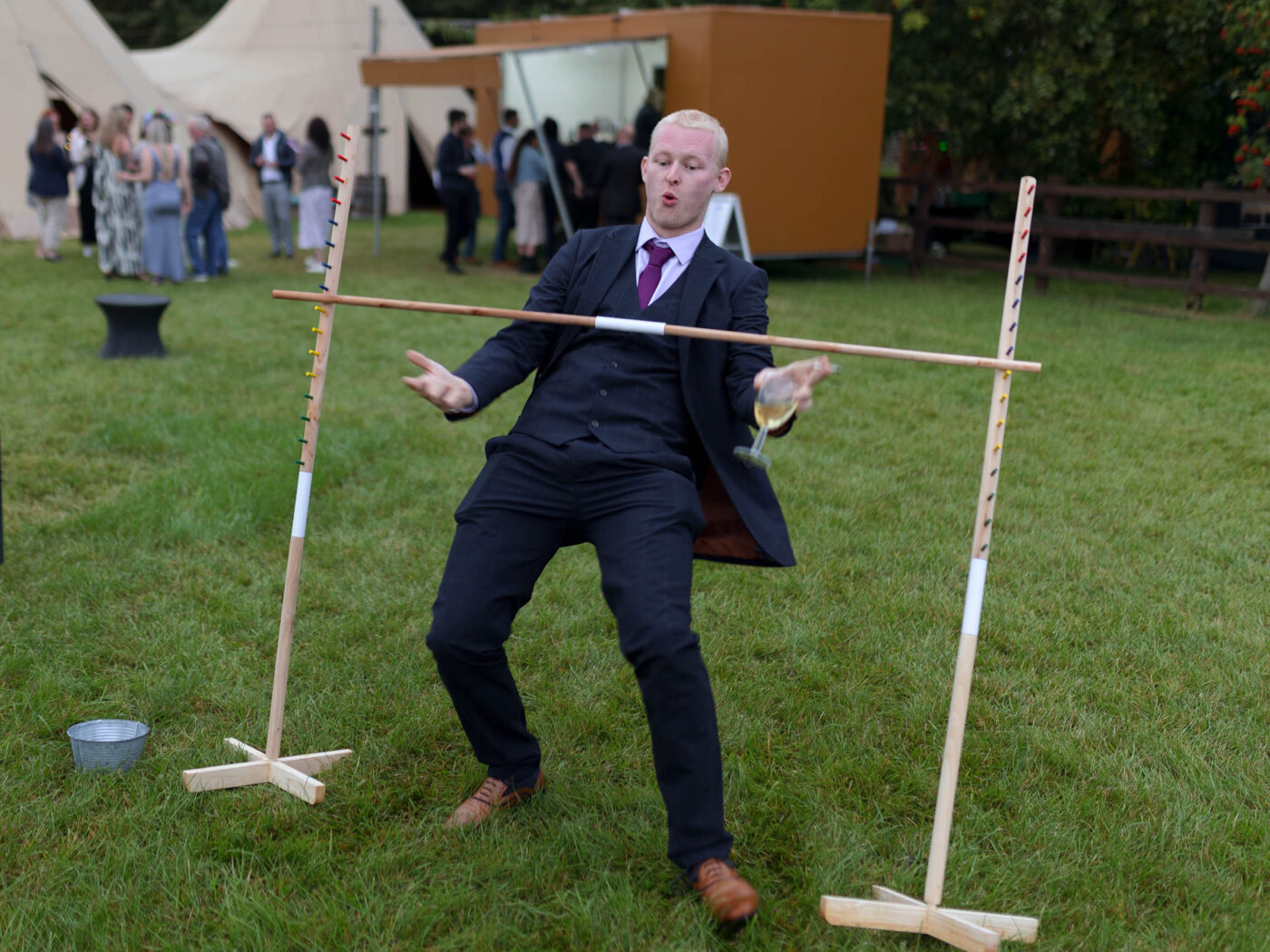 A man in a suit and a drink in his hand is leaning/falling backwards doing the limbo at a wedding party
