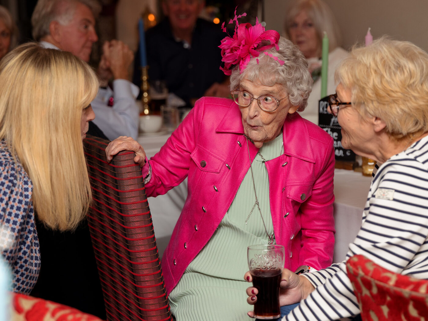 An elderly lady wearing a pink jacket and matching fascinator is looking at 2 wedding guests and has a cheeky naughty expression