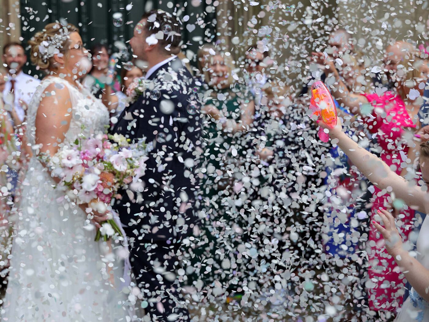 A storm of confetti hides a newlywed coupkle and their guests. to the corner of the frame is a young girls arm holding onto a brightly coloured bubble gun