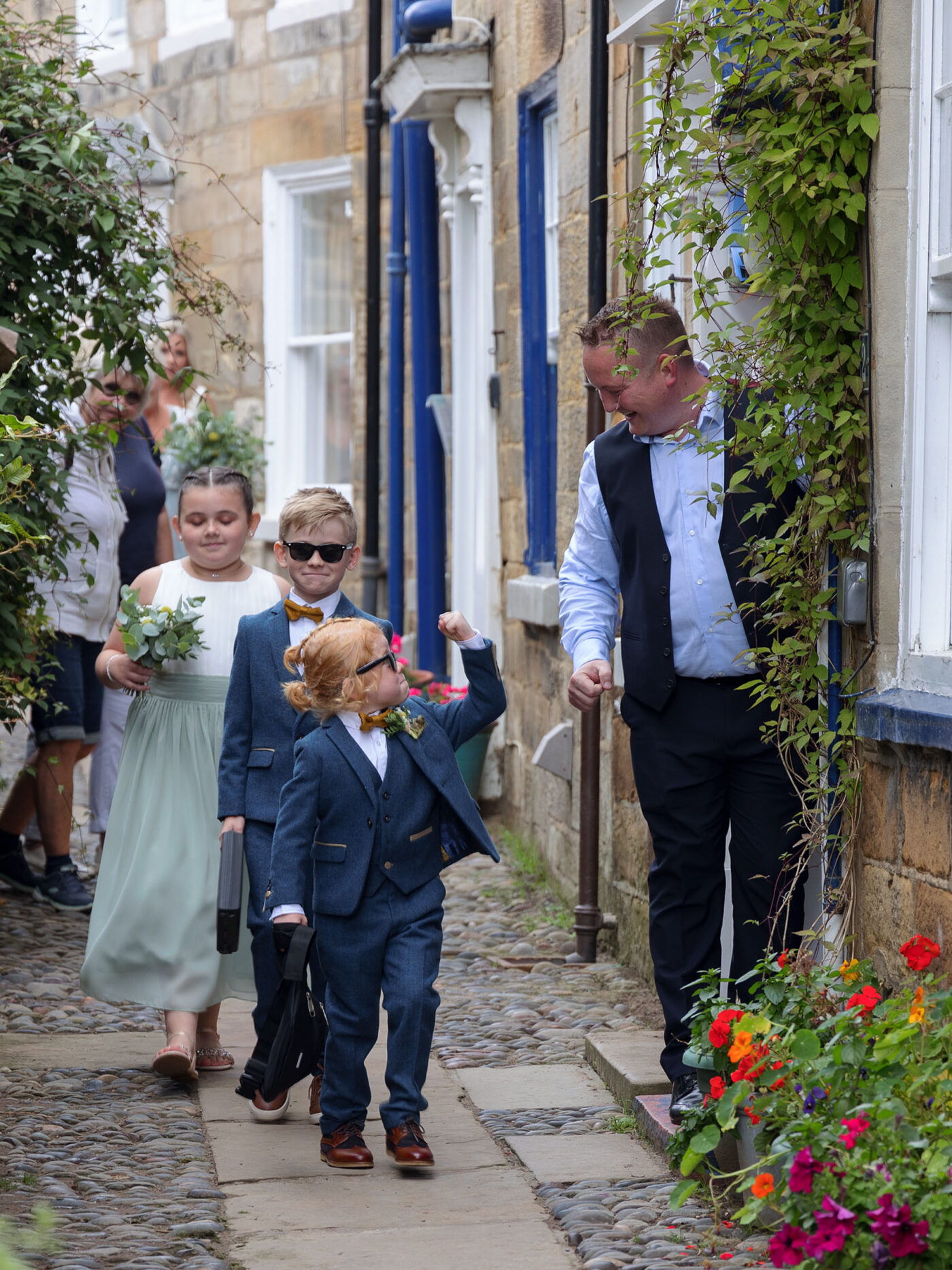2 Page boys and a young Bridesmaid are walking along a narrow street in Robin Hoods Bay, lined with colourful flowers and doorways. The boy at the front is waving his fist at a man coming out of one of the doors