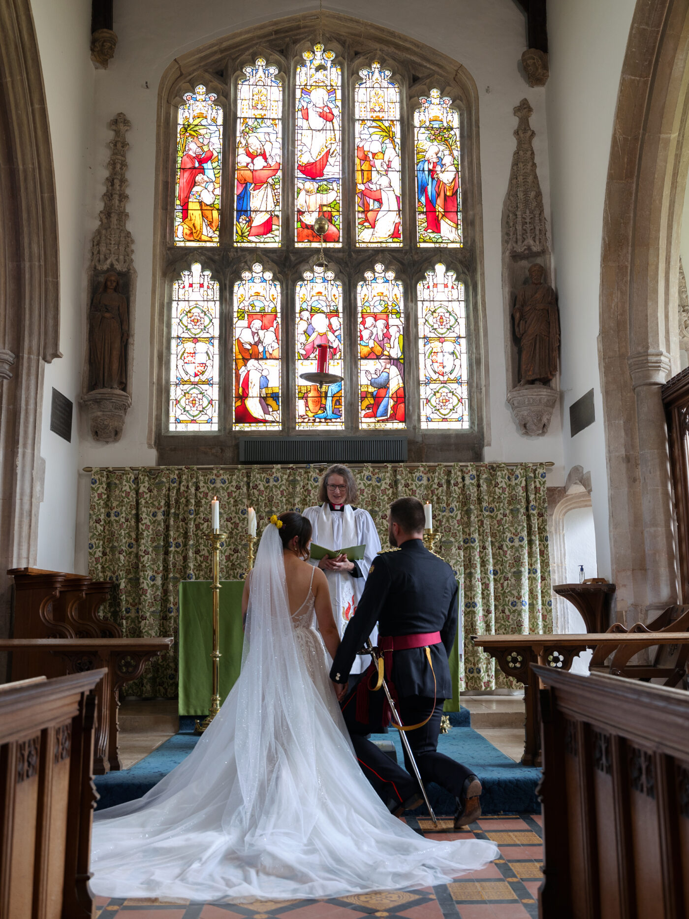 ride and groom kneel at the altar inside a church with stained glass windows during their ceremony.