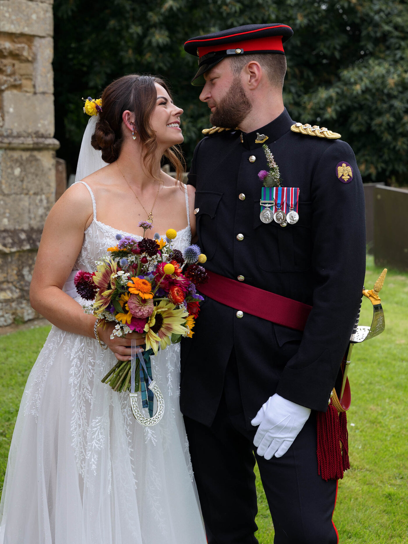 Bride holding a colorful bouquet smiles at a groom in formal military dress uniform during an outdoor wedding portrait at Harlaxton Church