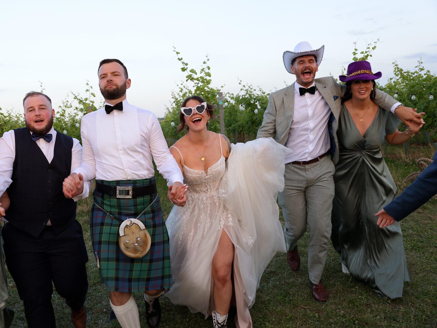Bride and groom run hand in hand across grass with their wedding party, some wearing hats and a kilt, all smiling and laughing.
