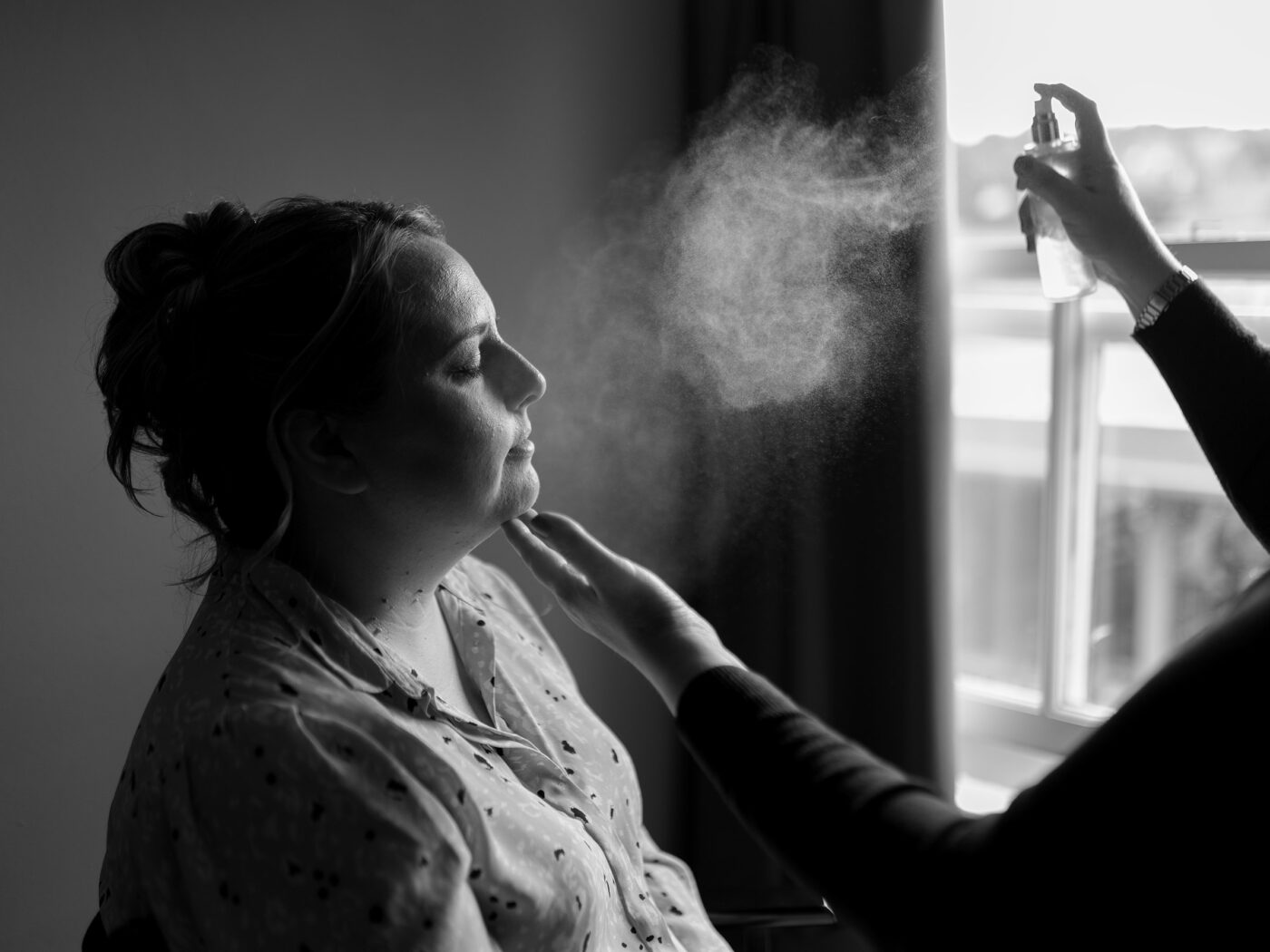 A woman sits in the window light having her make up done. The MUA is spraying her face with setting spray during Wedding morning prep