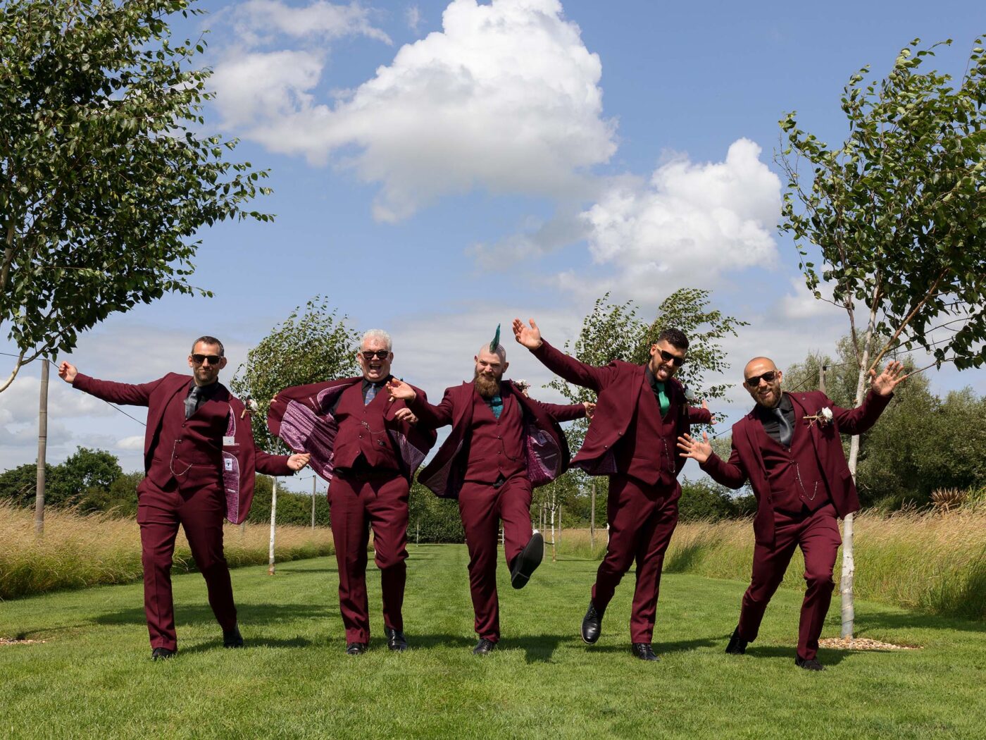 Five groomsmen in matching burgundy suits jump playfully in unison on a lawn under a blue sky.