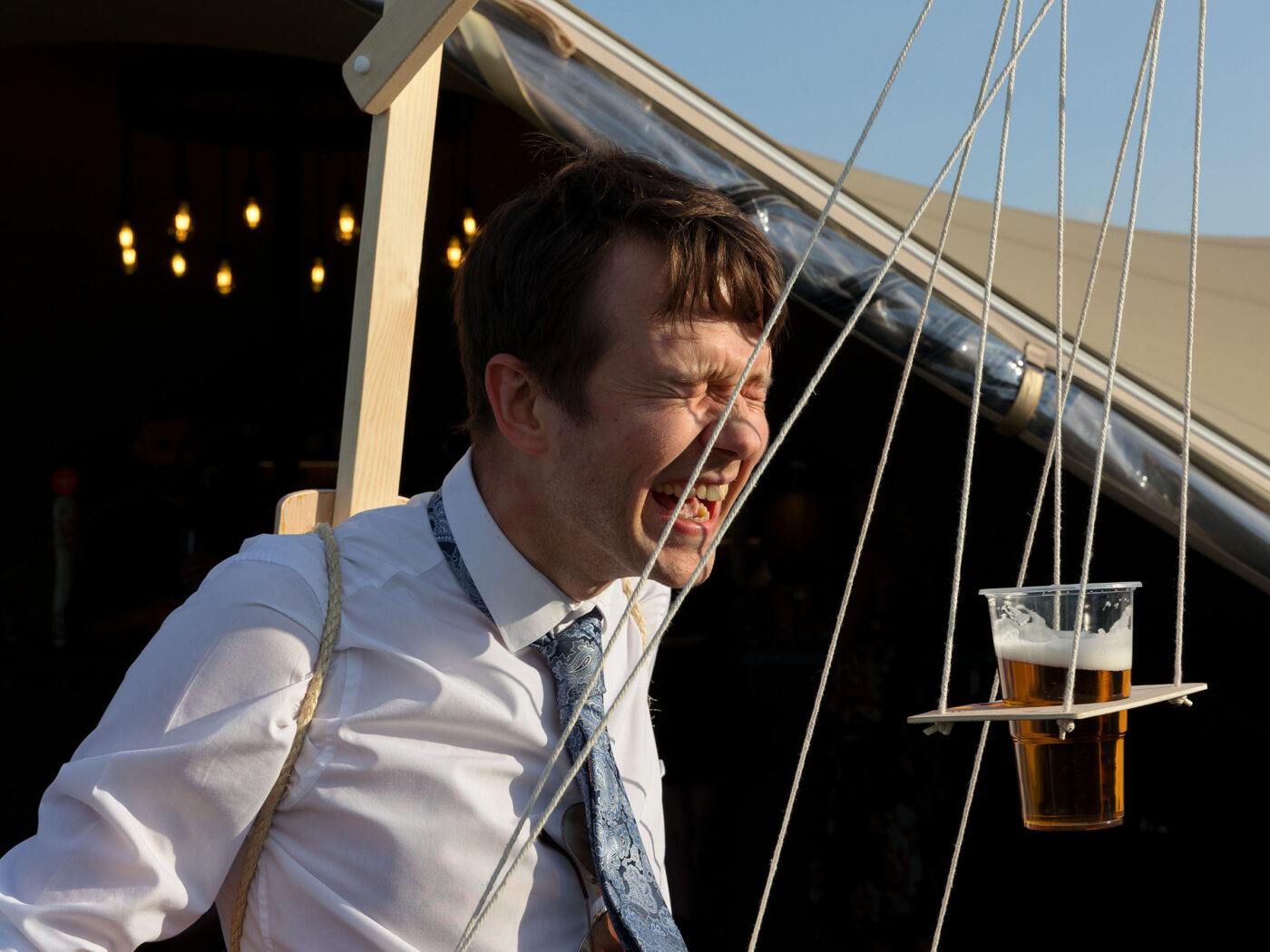 A smiling guest reacts as a tray holding pints of beer hangs suspended by ropes inf ront of him as part of a drinking game