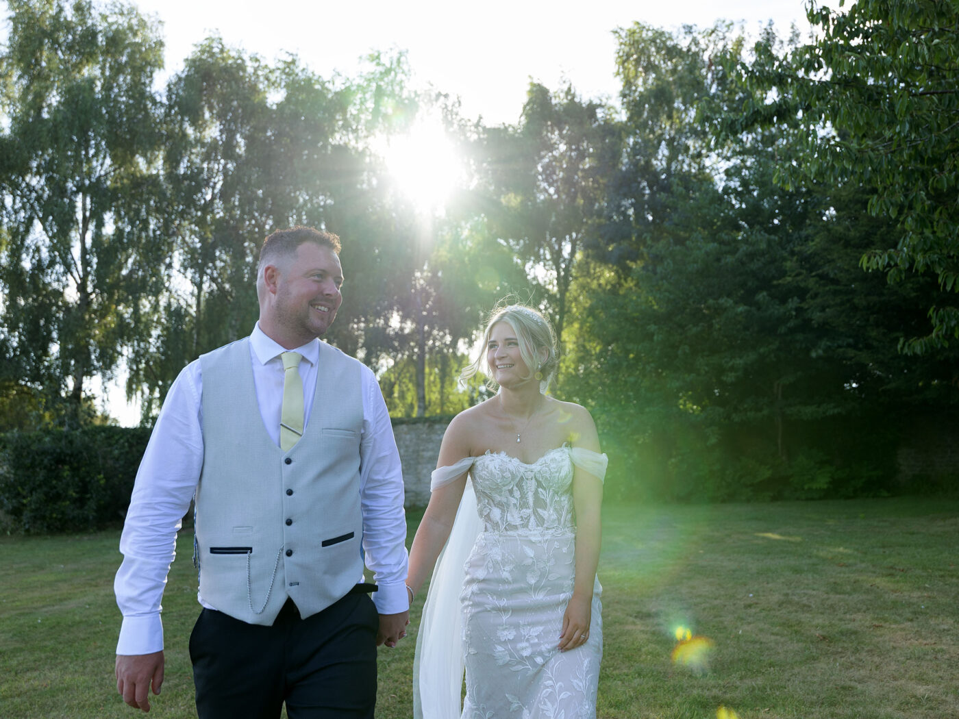 A bride and groom holding hands are walking towards the camera bathes in golden hour sunlight