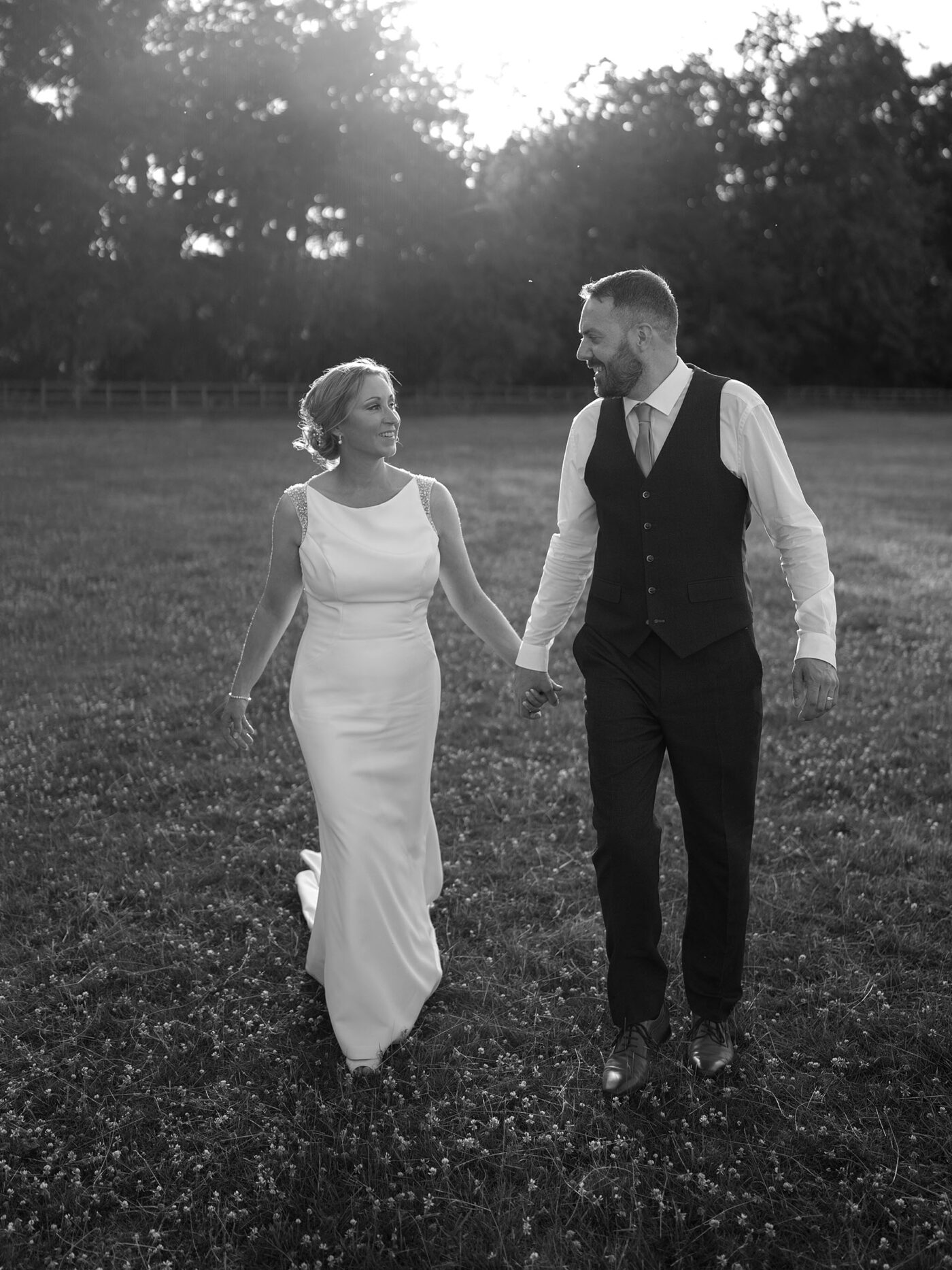 A bride and groom walking hand in hand through a grassy field during sunset; black-and-white wedding photo with soft natural light.