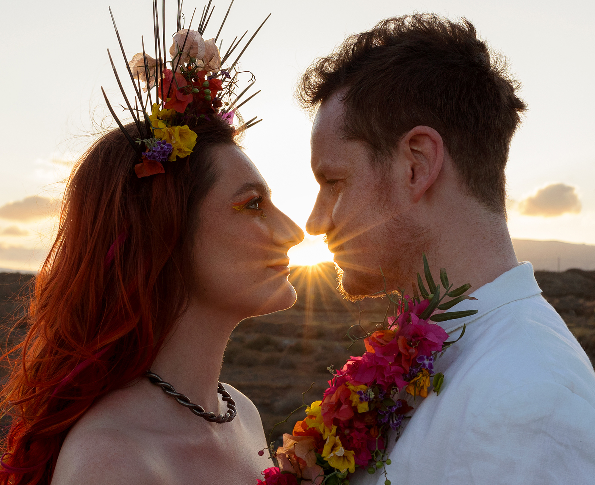 Alternative bride and groom stood facing each other looking into each others eyes. He is wearing a white suit with a floral lapel, she is wearing an ivory dress with puffy sleeves. The sun is setting behind creating a star burst between their faces