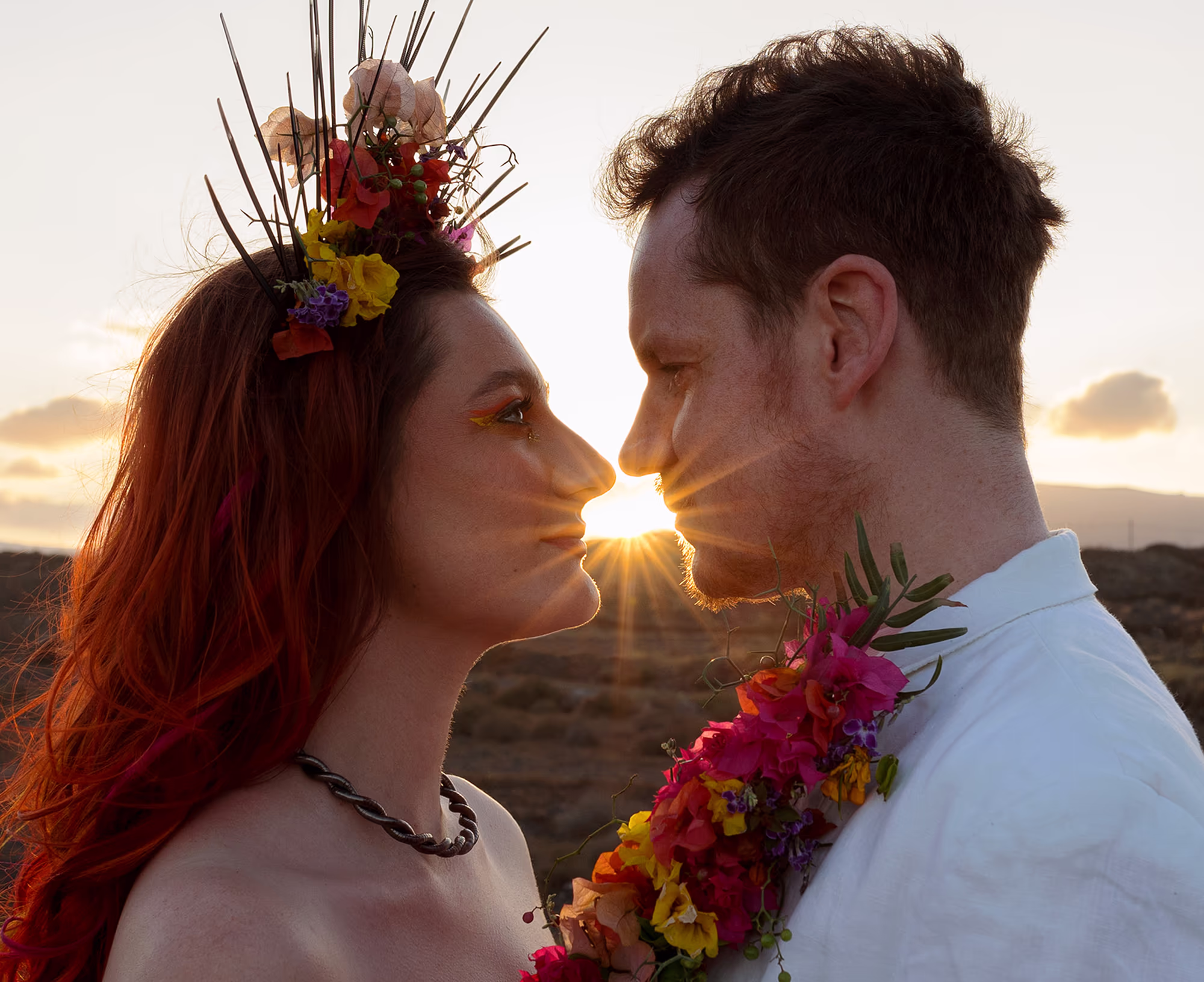 Bride and groom stood facing each other, the sun is setting behind them. The groom has his hand is his pocket