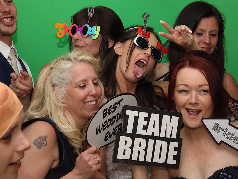 A group of women posing in a photo booth holding novelty props, including a “Team Bride” sign, smiling and laughing.