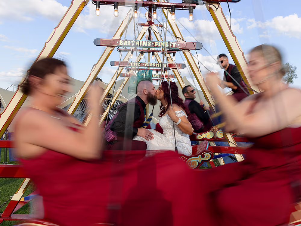Bride and groom kiss on a brightly lit fairground ride as bridesmaids in red dresses spin past in motion blur.