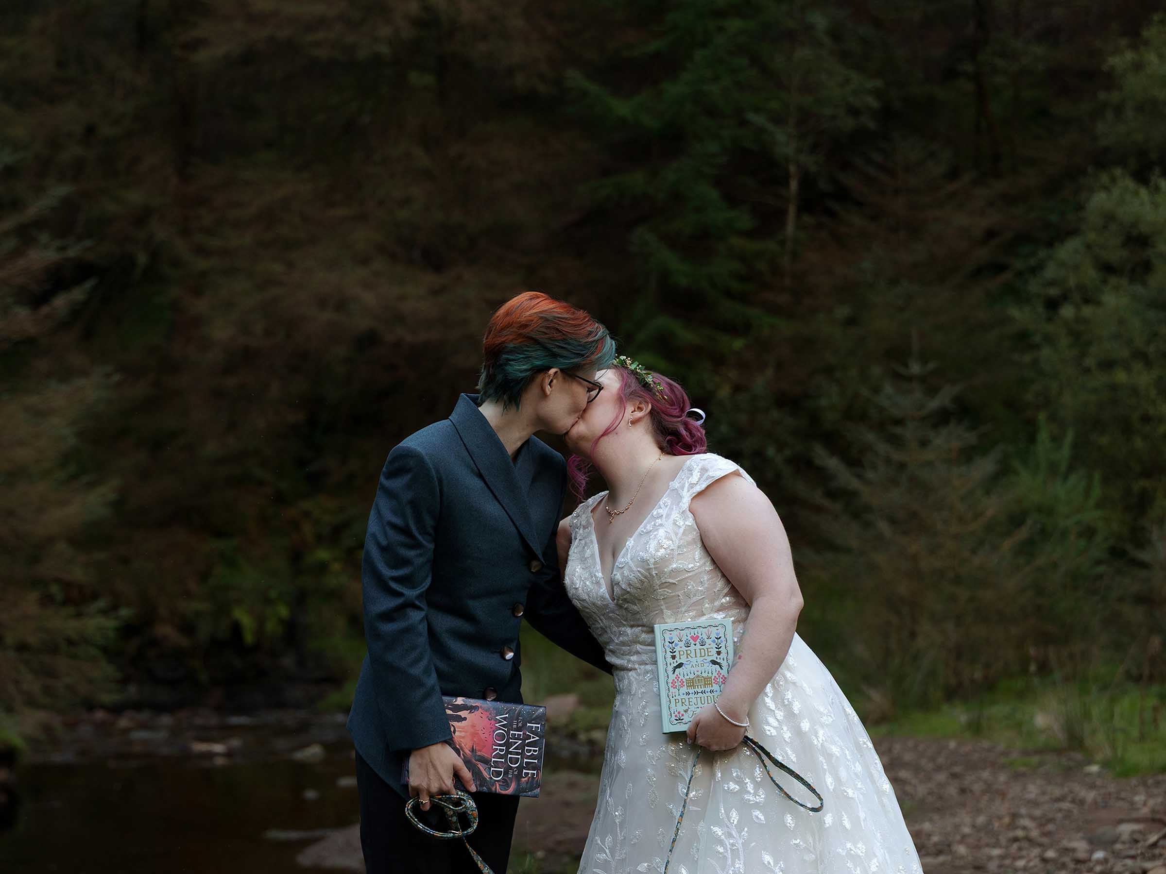 Newlyweds kiss in a forest setting holding books and dog leads