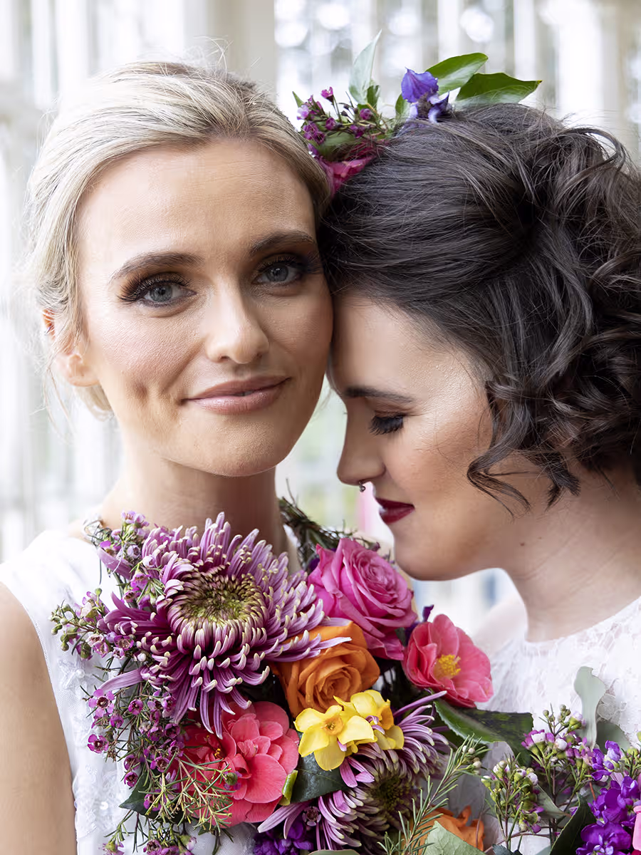 Wollaton Hall, Two brides with their faces close to each other. Tightly cropped photo of their faces. Blonde Bride looks at the camera, while the dark haired bride rests her forehead on her wifes cheek and closes her eyes. Both have brightly coloured flowers as a necklace and hair piece. taken by Pink Photographics Sleaford, Lincolnshire wedding Photographer
